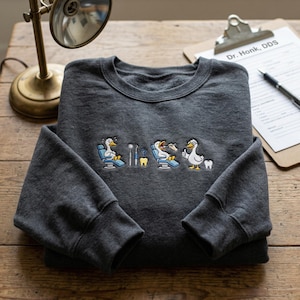 May include: A charcoal grey sweatshirt with a dental theme. The design features cartoon geese in a dentist's chair, dental tools, and teeth. The text "Dr. Honk, DDS" is visible on a clipboard.