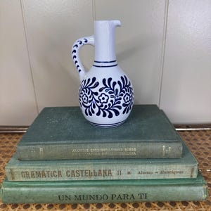 May include: A white ceramic pitcher with a blue floral design and a curved handle. The pitcher sits atop three vintage green books with Spanish text. The books are stacked on a woven surface.