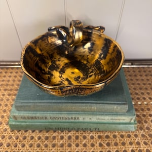 May include: A decorative bowl with a gold and black swirl design, featuring a handle. The bowl is placed on top of two vintage green books. The bowl's interior has a glossy finish.
