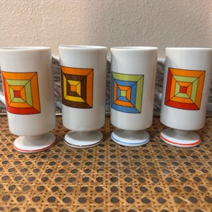 May include: Four white ceramic mugs with colorful geometric square designs. Each mug has a footed base with a colored ring. The geometric designs include orange, brown, green, and blue squares.