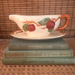 May include: Cream-colored gravy boat with a matching saucer. The boat features a handle and is decorated with red apples and green leaves on brown branches. The set is placed on top of three vintage books with Spanish titles.