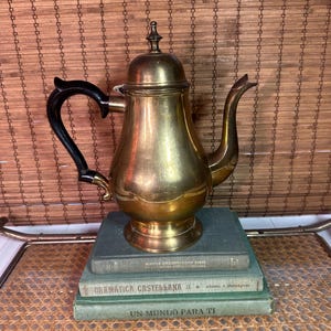 May include: A vintage brass tea or coffee pot with a black handle and spout. The pot sits atop a stack of three books with Spanish titles. The background is a woven bamboo blind.