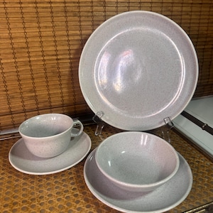 May include: A light gray speckled ceramic dish set, including a large plate, a smaller plate, a bowl, a teacup, and a saucer. The set is displayed on a woven surface, showcasing a minimalist aesthetic.