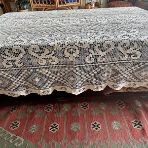 May include: A large, rectangular, cream-colored lace tablecloth with intricate floral and geometric patterns. The tablecloth is draped over a wooden table, with the edges hanging down. The tablecloth is placed on a red and green patterned rug.