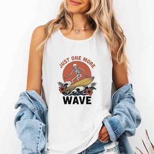 Puede incluir: Camiseta sin mangas blanca con un diseño gráfico de un esqueleto surfeando en una tabla amarilla, con el texto "JUST ONE MORE WAVE" en negro. El diseño incluye un sol rojo, olas y flores tropicales.