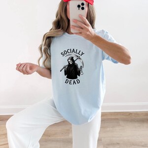 Może przedstawiać: Jasnoniebieski t-shirt z krótkim rękawem z czarno-białą grafiką przedstawiającą kostuchę trzymającą kosę i zegarek kieszonkowy. Napis "SOCIALLY DEAD" jest wydrukowany czcionką w łuk nad i pod centralną ilustracją przedstawiającą postać.