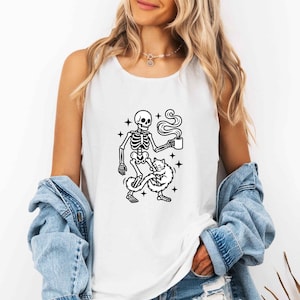 May include: White tank top featuring a black and white graphic of a skeleton holding a coffee cup, with a cat at its feet. The design includes stars and steam rising from the cup. A casual, fun design.