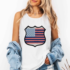 Può includere: Una canotta bianca senza maniche con una grafica centrale a forma di scudo. L'emblema presenta un design stilizzato della bandiera americana con stelle blu su campo azzurro e strisce orizzontali rosse e blu con una texture irregolare.