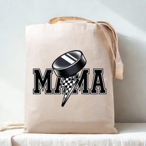 May include: A beige canvas tote bag featuring a black and white graphic print. The design displays the word MAMA in a bold varsity font, with a black hockey puck replacing the letter A and a checkered lightning bolt extending downward.