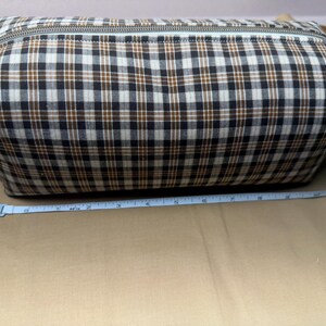 May include: A rectangular zippered pouch featuring a plaid pattern in black, white, and brown. A blue measuring tape is positioned along the base, indicating the pouch has a length of approximately 9 inches from end to end.