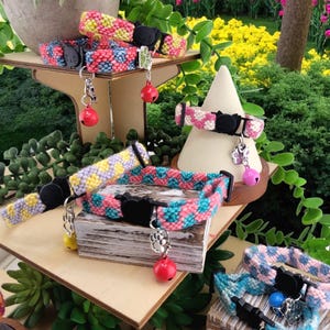Puede incluir: Una exhibición de coloridos collares para gatos con patrones tejidos en varios colores, incluyendo rosa, azul, amarillo y negro. Cada collar presenta una hebilla negra con forma de gato y un pequeño dije con una campana. Los collares se exhiben en soportes de madera.