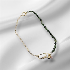 May include: A gold necklace with a chain and a strand of green beads. The necklace has a toggle clasp and a decorative clasp with a small heart and pearl charm. The necklace is displayed on a white surface.