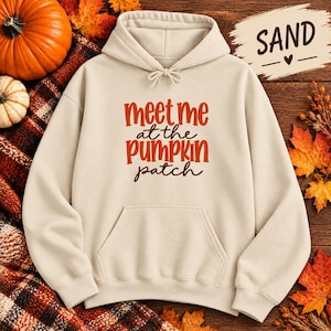 May include: A sand-colored hooded sweatshirt featuring the text "meet me at the pumpkin patch" printed in orange and dark brown lettering. The hoodie includes a front kangaroo pocket, long sleeves, and a drawstring hood with matching sand-colored cords.