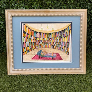 May include: Watercolor painting of a library scene. A person is lying on a blue chaise lounge on a red rug, reading a book. The library is filled with colorful books on shelves. The painting is framed in a light-colored wooden frame.