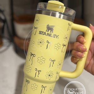 Pu&ograve; includere: Tazza da viaggio in acciaio inossidabile gialla con un motivo di margherite e fiocchi in bianco e nero. La tazza ha un manico e un coperchio con una cannuccia. La tazza ha la scritta "STANLEY" stampata su di essa.