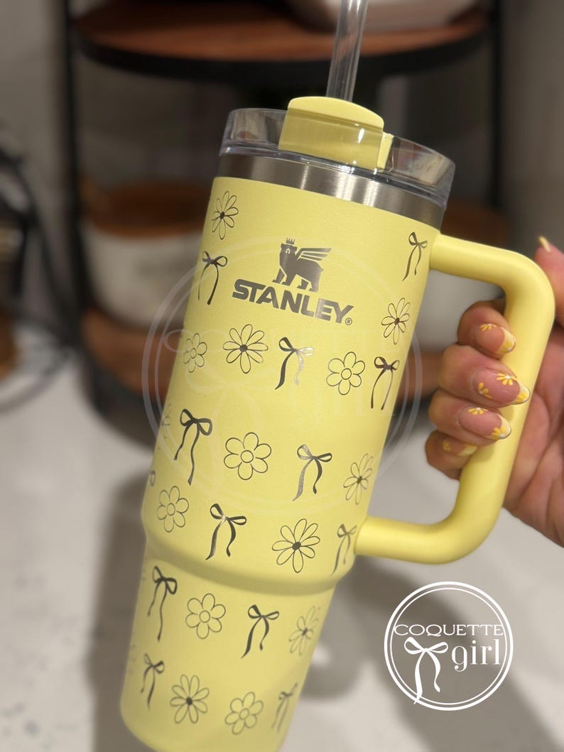 Pu&ograve; includere: Tazza da viaggio in acciaio inossidabile gialla con un motivo di margherite e fiocchi in bianco e nero. La tazza ha un manico e un coperchio con una cannuccia. La tazza ha la scritta "STANLEY" stampata su di essa.