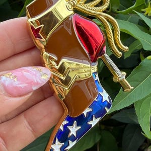 May include: A high-heeled boot brooch featuring a brown shaft, a red heel, and a blue toe section decorated with white stars. The design includes gold-tone accents, a gold-tone rope detail at the top, and a gold-tone emblem on the side.