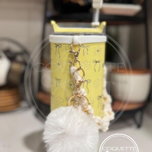 Pu&ograve; includere: Un bicchiere giallo con un coperchio bianco e un motivo floreale bianco e giallo. Il bicchiere ha un portachiavi a pompon bianco attaccato a una catena dorata e un cinturino in crochet bianco. Il portachiavi ha anche un anello per chiavi dorato. Il bicchiere &egrave; appoggiato su una superficie di legno. Il testo "Coquette girl" &egrave; visibile in basso a destra dell'immagine.