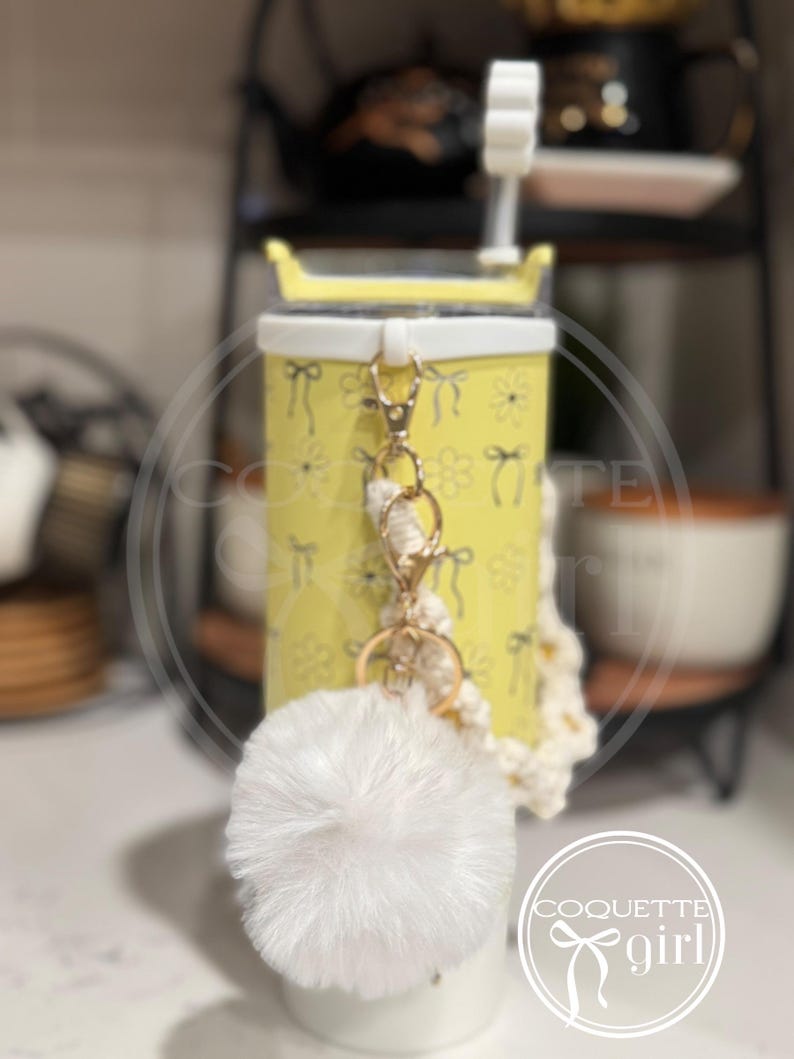 Pu&ograve; includere: Un bicchiere giallo con un coperchio bianco e un motivo floreale bianco e giallo. Il bicchiere ha un portachiavi a pompon bianco attaccato a una catena dorata e un cinturino in crochet bianco. Il portachiavi ha anche un anello per chiavi dorato. Il bicchiere &egrave; appoggiato su una superficie di legno. Il testo "Coquette girl" &egrave; visibile in basso a destra dell'immagine.