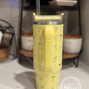 Pu&ograve; includere: Un bicchiere in acciaio inossidabile giallo con un coperchio trasparente e una cannuccia. Il bicchiere ha un motivo di fiocchi grigi e fiori bianchi. Il bicchiere &egrave; appoggiato su un bancone bianco.