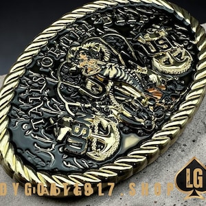 May include: An oval-shaped belt buckle with a gold rope-like border. The center features a black background with gold-colored raised relief details, including anchors, a ship, and the text "USN". The buckle has the text "OF THE UNITED STATES" and "LG" on it.