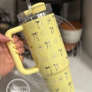 Pu&ograve; includere: Un bicchiere termico in acciaio inossidabile giallo con una maniglia nera e una cannuccia. Il bicchiere ha un motivo di fiocchi neri e fiori. Il bicchiere &egrave; etichettato "Coquette girl" con un fiocco nero.