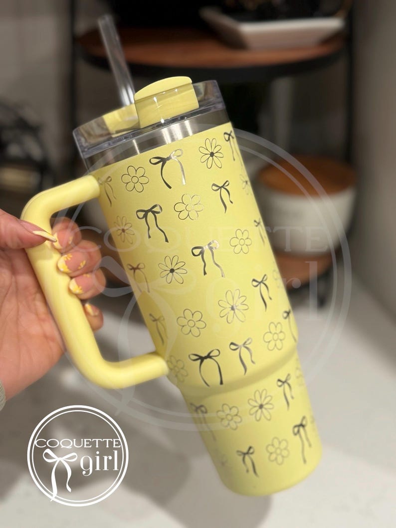Pu&ograve; includere: Un bicchiere termico in acciaio inossidabile giallo con una maniglia nera e una cannuccia. Il bicchiere ha un motivo di fiocchi neri e fiori. Il bicchiere &egrave; etichettato "Coquette girl" con un fiocco nero.