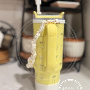 Pu&ograve; includere: Un bicchiere giallo con un coperchio bianco e un manico in corda bianco e giallo. Il bicchiere ha un motivo di fiocchi e fiori. Il bicchiere &egrave; appoggiato su una superficie bianca. Il testo "Coquette girl" &egrave; visibile in basso a destra dell'immagine.