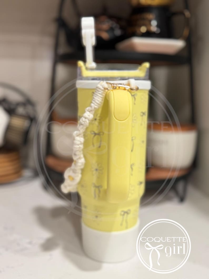 Pu&ograve; includere: Un bicchiere giallo con un coperchio bianco e un manico in corda bianco e giallo. Il bicchiere ha un motivo di fiocchi e fiori. Il bicchiere &egrave; appoggiato su una superficie bianca. Il testo "Coquette girl" &egrave; visibile in basso a destra dell'immagine.