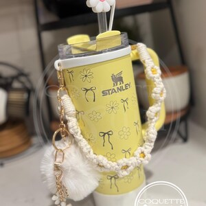 Pu&ograve; includere: Un bicchiere termico in acciaio inossidabile giallo con un tappo di cannuccia a forma di margherita bianca, una tracolla in crochet bianco e un portachiavi a pompon bianco. Il bicchiere ha un motivo a margherite e fiocchi e il testo "STANLEY" e "COQUETTE girl".