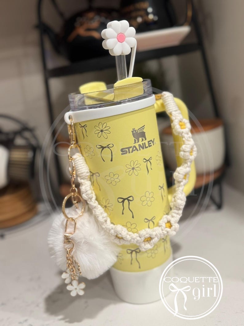 Pu&ograve; includere: Un bicchiere termico in acciaio inossidabile giallo con un tappo di cannuccia a forma di margherita bianca, una tracolla in crochet bianco e un portachiavi a pompon bianco. Il bicchiere ha un motivo a margherite e fiocchi e il testo "STANLEY" e "COQUETTE girl".