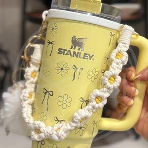 Pu&ograve; includere: Tazza da viaggio Stanley gialla con un motivo a margherita bianca e un manico a crochet di margherita bianca. La tazza ha un coperchio bianco con una cannuccia di plastica trasparente e un topper di cannuccia a margherita bianca. La tazza ha il testo "STANLEY" sul davanti.