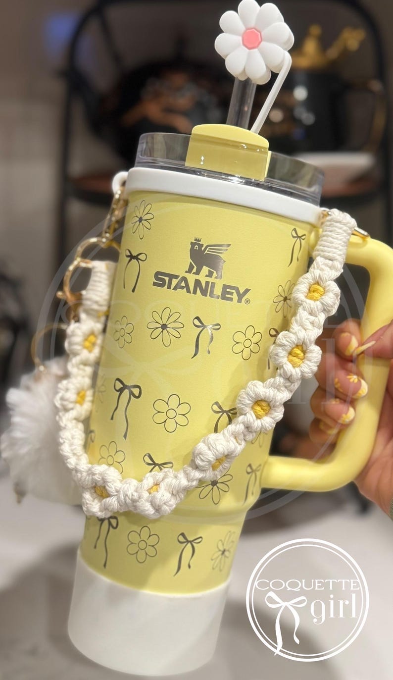 Pu&ograve; includere: Tazza da viaggio Stanley gialla con un motivo a margherita bianca e un manico a crochet di margherita bianca. La tazza ha un coperchio bianco con una cannuccia di plastica trasparente e un topper di cannuccia a margherita bianca. La tazza ha il testo "STANLEY" sul davanti.
