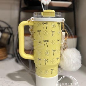 Pu&ograve; includere: Un bicchiere giallo con un coperchio bianco e un motivo bianco e nero di fiocchi e fiori. Il bicchiere ha un manico e una base bianca. Il bicchiere ha un ciondolo bianco con il testo "Coquette girl" su di esso.