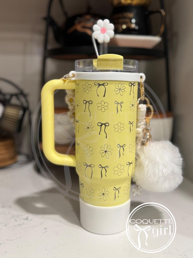Pu&ograve; includere: Un bicchiere giallo con un coperchio bianco e un motivo bianco e nero di fiocchi e fiori. Il bicchiere ha un manico e una base bianca. Il bicchiere ha un ciondolo bianco con il testo "Coquette girl" su di esso.