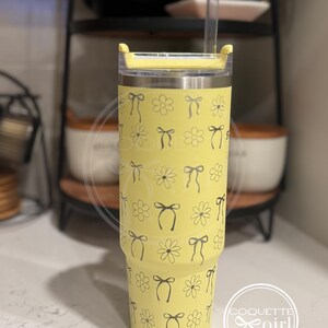 Pu&ograve; includere: Un bicchiere in acciaio inossidabile giallo con un motivo di margherite e fiocchi neri e bianchi. Il bicchiere ha un coperchio e una cannuccia.