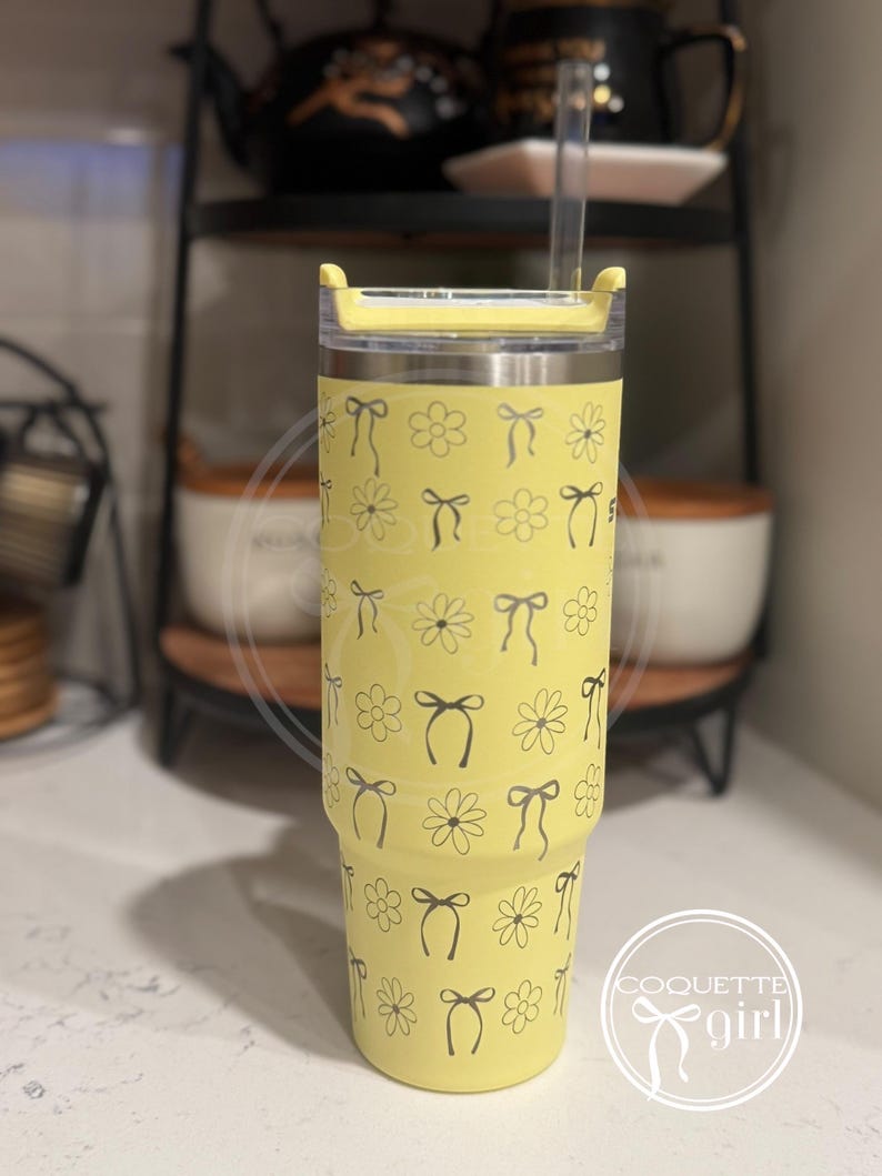 Pu&ograve; includere: Un bicchiere in acciaio inossidabile giallo con un motivo di margherite e fiocchi neri e bianchi. Il bicchiere ha un coperchio e una cannuccia.