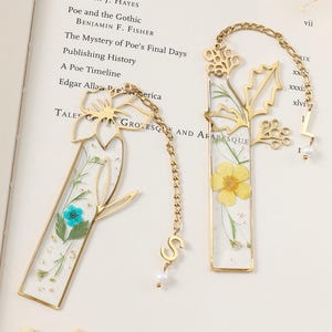 May include: Two gold-coloured metal bookmarks with floral designs. One features a blue flower, the other a yellow flower, both encased in clear resin. Each has a chain with a letter charm and a small pearl.