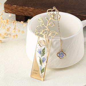 Peut inclure: Un marque-page en métal doré avec un motif floral découpé sur le dessus et une section en résine transparente contenant des fleurs bleues et des feuilles vertes pressées. Le texte "Rooted in hope" est imprimé sur le métal, et une petite breloque en forme de trèfle avec une fleur bleue est suspendue à une fine chaîne dorée.