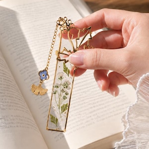 Könnte beinhalten: Ein rechteckiges Lesezeichen mit gepressten weißen Blumen und grünem Blattwerk in klarem Harz mit einem goldfarbenen Metallrahmen. An der Oberseite befindet sich eine goldene Kette mit einem kleinen blauen Blumenanhänger und einem goldenen Ginkgoblatt.