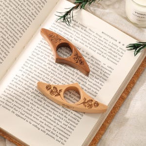 May include: Two wooden page holders with a central circular opening for a thumb, featuring engraved floral designs. One holder is crafted from a darker, reddish-brown wood, while the other is a lighter, natural beige wood, both resting on an open book.