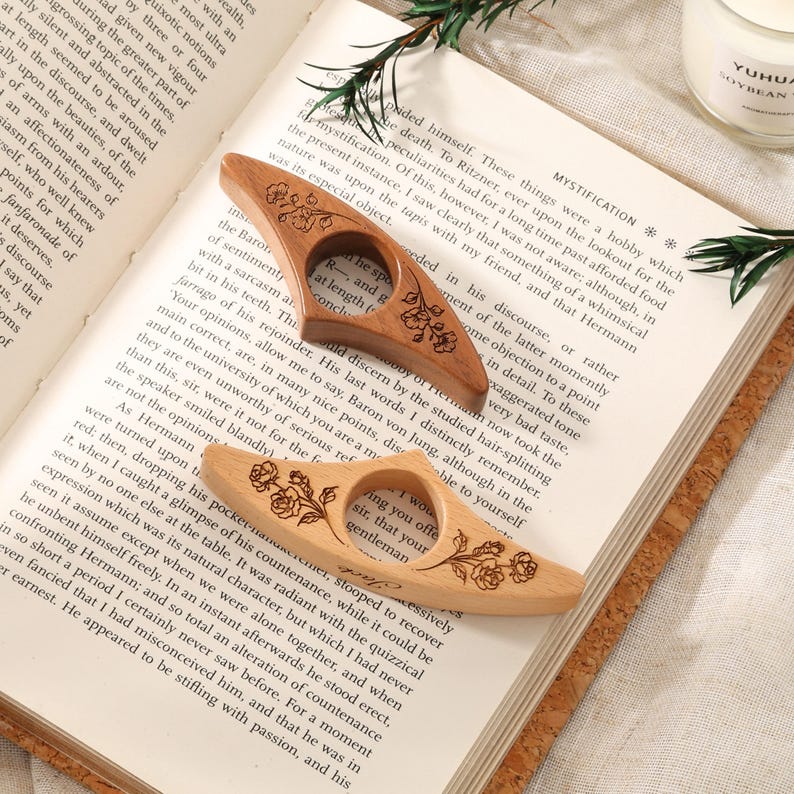 May include: Two wooden page holders with a central circular opening for a thumb, featuring engraved floral designs. One holder is crafted from a darker, reddish-brown wood, while the other is a lighter, natural beige wood, both resting on an open book.