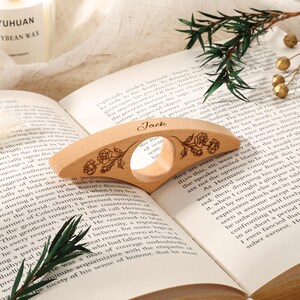 May include: A light-colored wooden page holder with a central circular cutout, featuring the engraved name Jack and floral rose designs on both sides. The object is positioned horizontally across the open pages of a printed book.