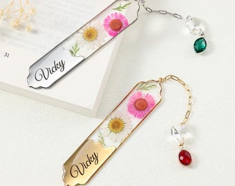 May include: Two personalized bookmarks with embedded flowers and the name "Vicky". One bookmark is silver with a green gem, the other is gold with a red gem. Both have a chain and crystal accents.