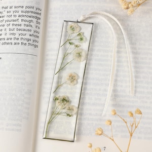 May include: A clear resin bookmark with dried white flowers and green stems encased within. A white ribbon is attached. The bookmark is rectangular and is lying on an open book.