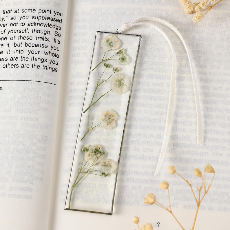 May include: A clear resin bookmark with dried white flowers and green stems encased within. A white ribbon is attached. The bookmark is rectangular and is lying on an open book.