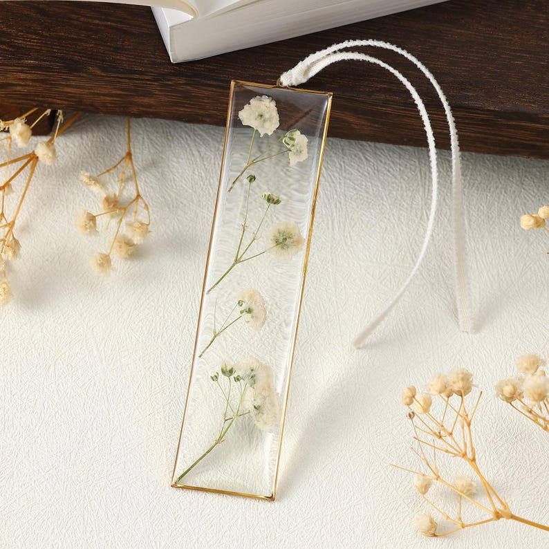 May include: A rectangular bookmark with a gold-coloured frame encases delicate white flowers in clear resin. A white cord is attached at the top. The bookmark is set against a white surface with dried flowers.