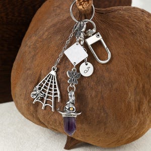 Puede incluir: Llavero plateado con múltiples colgantes. Incluye una telaraña con una araña, una mariposa, un cuadrado, un colgante redondo con la letra "J" y un sombrero de bruja con un cristal morado. El llavero tiene un mosquetón.