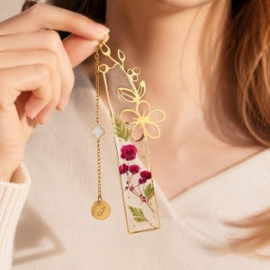 May include: A rectangular bookmark featuring clear resin embedded with pressed magenta flowers and green foliage, framed in gold metal with a floral cutout top. A gold chain with a white clover charm and a round pendant engraved with the letter J.