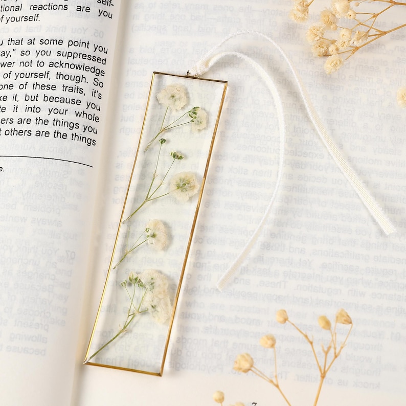 May include: A rectangular bookmark with a gold-coloured frame encases dried white flowers. A white ribbon is attached at the top. The bookmark is set against an open book.